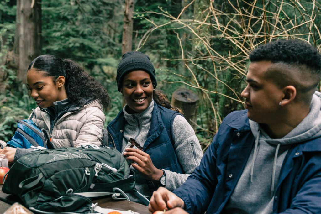 Three friends enjoying a break during a hiking trip in a lush forest, sharing smiles and snacks.
